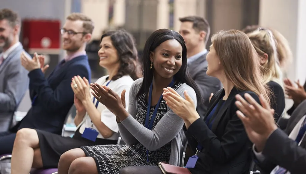People sitting down in chairs, smiling, and clapping at an event.