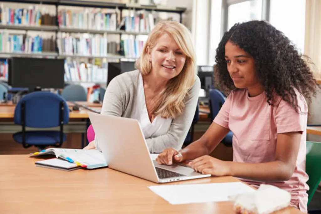 Smiling female teacher and student sitting at a desk in a classroom setting looking at a laptop.